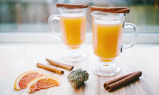 Apple cider infused with cannabis on a wooden table with cinnamon sticks, orange wedges, and cannabis flower