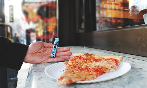 Hand holding cannabis vaporizer next to slice of pizza outside Joe's Pizza in NYC