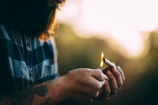 Man in blue plaid shirt lighting cannabis joint outside at sunset