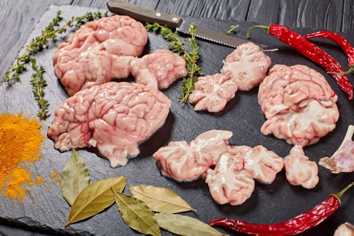 uncooked beef or calf's brains slices prepared to be cooked with spices, garlic, dried chillies, and herbs on black stone plate on wooden table, view from above, close-up