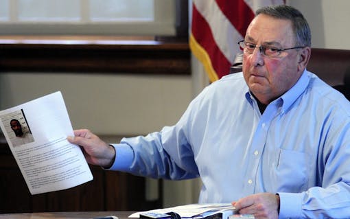 Maine Gov. Paul LePage holds up news release with a booking mug shot from a three-ring binder of news releases and articles about drug arrests during a meeting with reporters on Friday, Aug. 26, 2016, in the State House Cabinet room in Augusta, Maine. (Joe Phelan /The Kennebec Journal via AP)