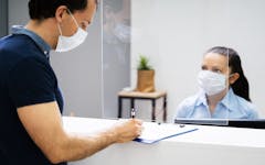 A masked patient signs in at a doctors office front desk. Stock photo.