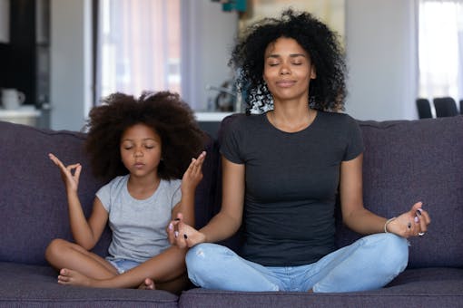 Mom and daughter meditate, sit with crossed legs, and smile on couch