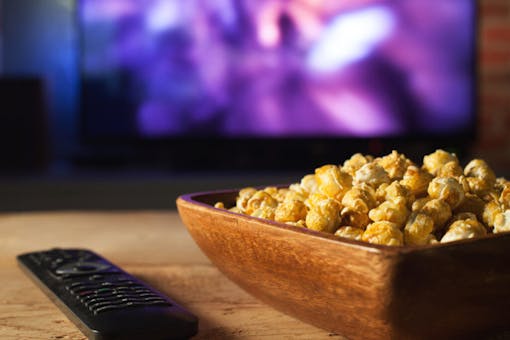A wooden bowl of popcorn and the TV remote in the background the TV works. Evening cozy watching a movie or TV series at home.