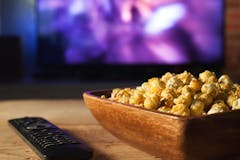 A wooden bowl of popcorn and the TV remote in the background the TV works. Evening cozy watching a movie or TV series at home.