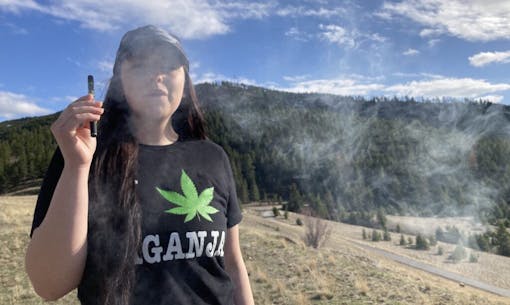 young woman with long brown hair and black t shirt reading Laganja with a green pot leaf exhales from a vape pen in front of a large stand of pine trees