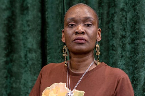 an older Black woman with large earrings, short hair, and a brown t shirt stands in front of a green curtain