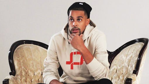 a Black man in a white sweatshirt and black beanie sits on a chair with a hand on his chin against a white interior wall