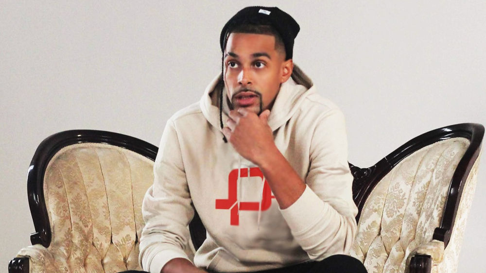 A black man in a white sweatshirt and black beanie sits in a chair with a hand on his chin against a white interior wall