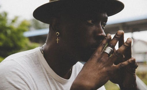 a Black man with a white t shirt and a wide brimmed hat steeples his hands in front of his face and looks into distance