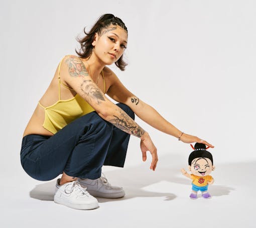 young woman with tattoos on her arm squats next to a six inch high figurine that she created against a white backdrop