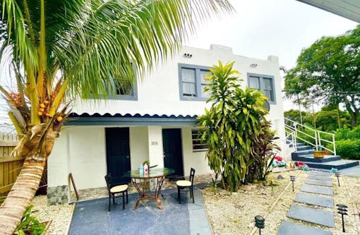 back patio and back of house with palm trees and a glass table with two chairs. to the right, a path leads to stairs that wrap around to front of house
