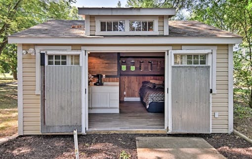 looking into tiny home from exterior. big farm doors have slid open to reveal quaint kitchen and big comfy bed