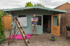 Older man with beard proudly standing beside his freshly painted garden shed.