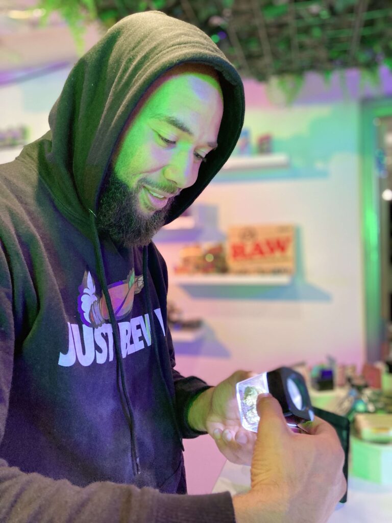 Calvin wears a black hoodie and smiles as he inspects the weed jar