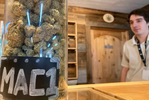 big deli style jar of MAC1 weed on counter with white man standing to the right