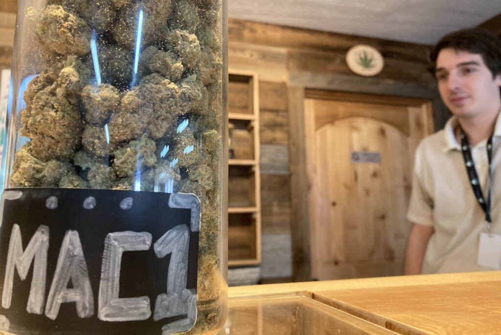 Large deli jar of MAC1 weed on counter with white man standing to the right