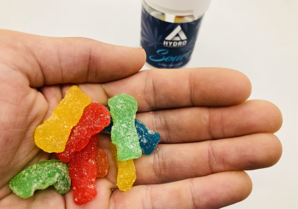 Outstretched hand holding multi colored bite sized gummies, with gummies in the background, against a white background