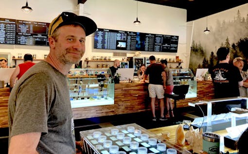 smiling man in front of long wooden sales counter with glass cases displaying cannabis products