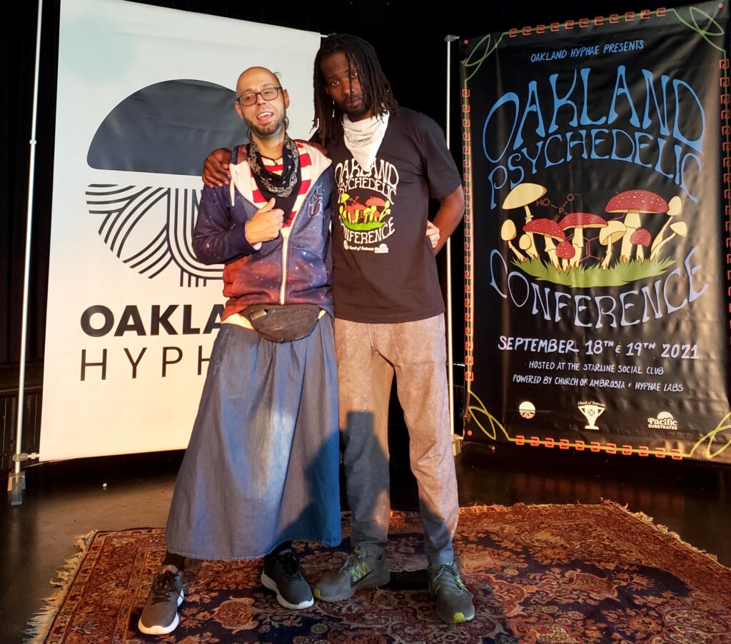 Two men, one in a blue skirt, stand in front of the Oakland Hyphae sign and the Psych Conference sign