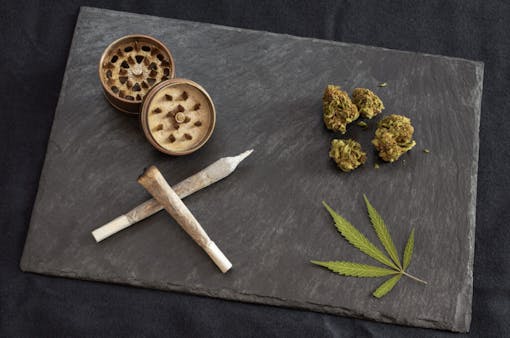 Marijuana joints ready to smoke, high quality cannabis buds, grinder and marijuana leaf in black stone tray. Gourmet weed on dark background.