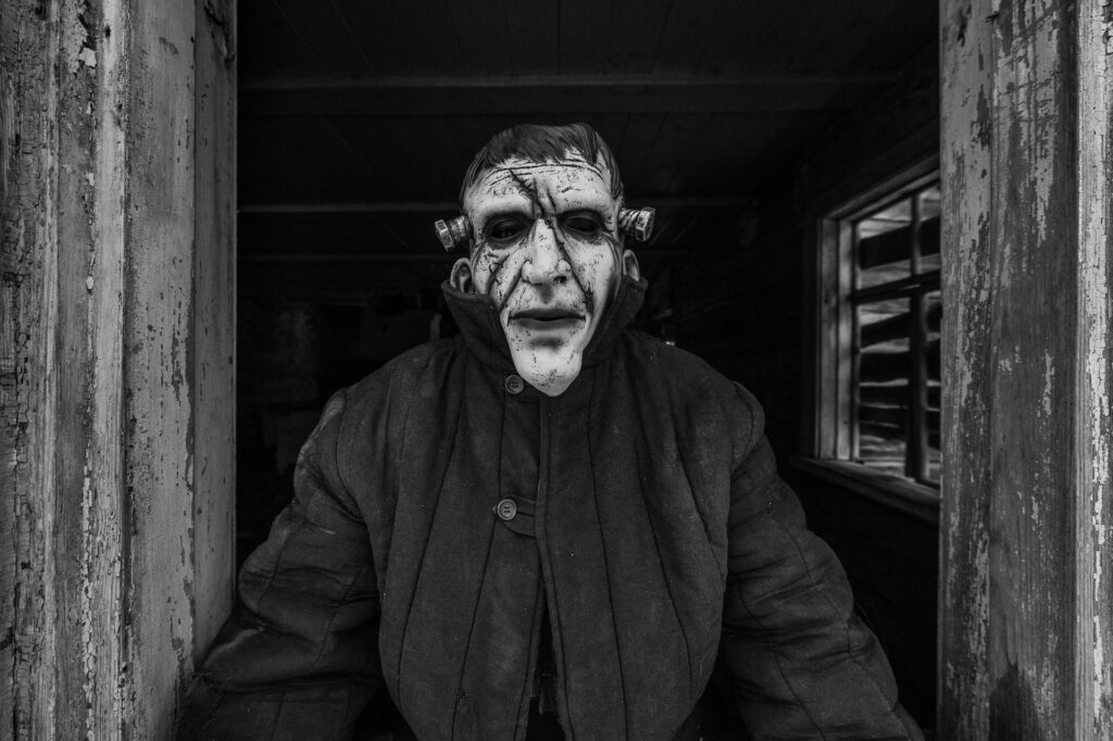 Unidentified person in spooky terrible latex mask looking out of old wooden haunted house window.