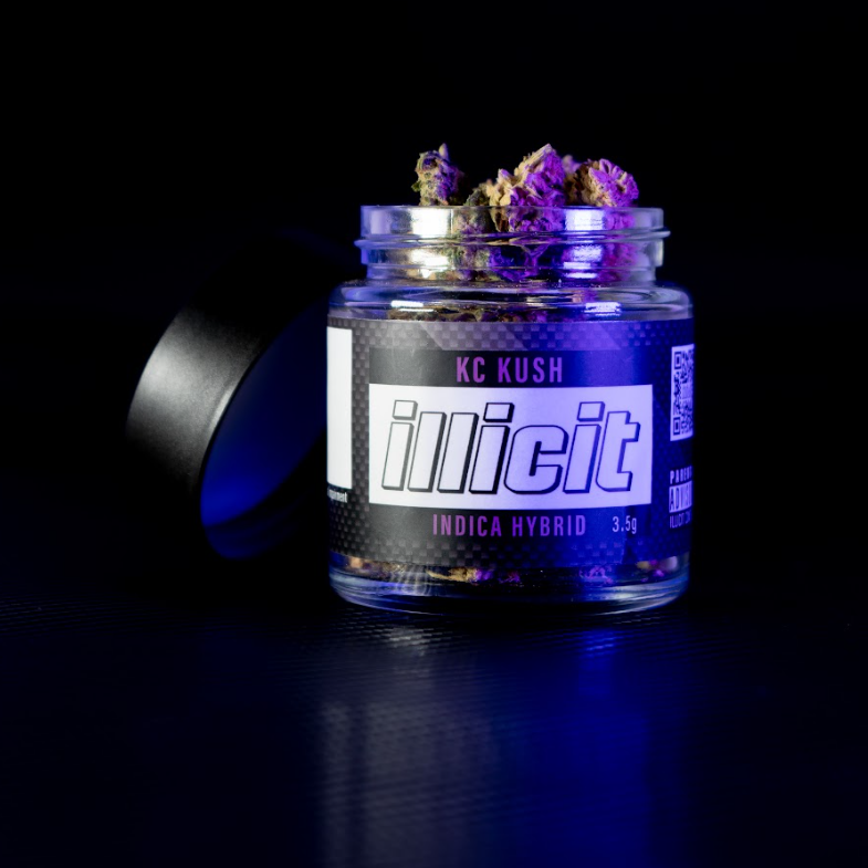 Photo of Illicit KC Kush on a black background.  The lid is removed from the jar and shows flower and label reads