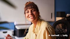 smiling woman with bangs in a yellow shire in a home office. bottom right corner features Leaflywell and Leafly's logos in white