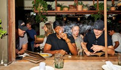 four people at a high counter inside, with many plants behind them. a man in center of frame is exhaling smoke from a joint.