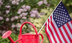 photo of American flag sticking out of red watering pail with out-of-focus flowering bush in the background
