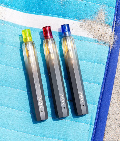 Three AiroX 1g vapes sitting in a line, laying down on a light blue beach mat with sand dusting the side