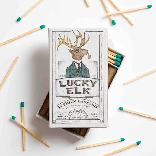 A box of Lucky Elk THCA pre-rolled joints with the box top sitting askew on top of the open box over a white background with bright green-blue matches scattered around