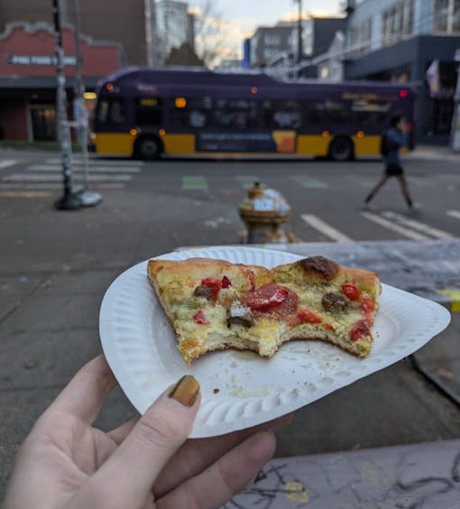 A photo of a woman's hand holding a paper plate with a half-eaten pizza slice on it from Hot Mama's, outside of their Capitol Hill Seattle location. One of our picks for the best munchies in Seattle.